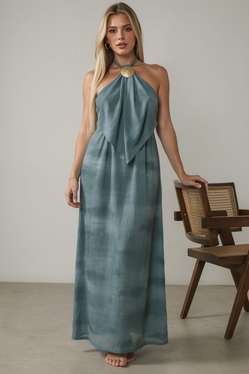 Woman wearing a long blue dress standing next to a wooden chair against a neutral background